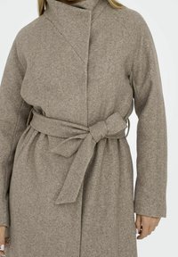 Béžový kabát z vlněné směsi s vysokým límcem, dlouhými rukávy a páskem na stažení. Má hladkou texturu a štíhlou siluetu.