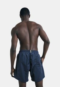 Pantaloni da bagno navy realizzati in tessuto leggero con vita elastica, tasca laterale e una texture liscia. Ideali per l'abbigliamento da spiaggia.