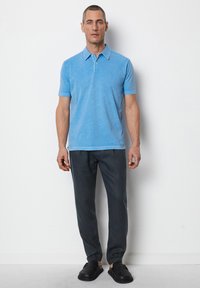 Hellblaues Poloshirt mit klassischem Kragen und zwei Knöpfen, kombiniert mit dunkelgrauen Hosen und schwarzen Slippern.