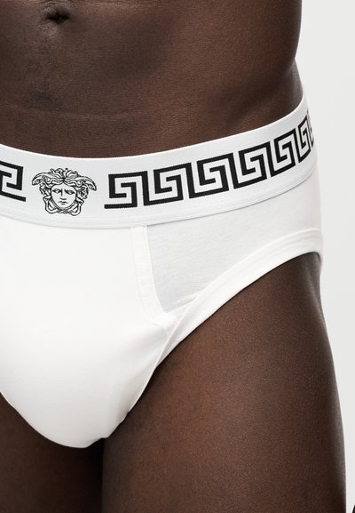 Gros plan sur une personne portant un slip blanc avec un motif Greca noir et un logo Méduse sur la ceinture, vu de la taille vers le bas.