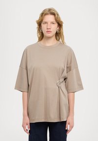 T-shirt beige oversize realizzato in cotone morbido, con scollo rotondo e un accento a forma di anello argento sul lato. Texture liscia, vestibilità comoda.
