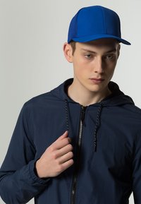 Kék sapka, szilárd elülső résszel és hálós oldalakkal, egy személyen, aki tengerészkék dzsekit visel, cipzárral és húzózsinórokkal. Sima anyag, laza fit.