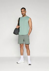 Vyriški sportiniai drabužiai apima šviesiai žalią rankovių neturintį viršutinį drabužį ir pilką šortus, derinamus su juoda rankine. Baltos sportinės bateliai ir kulkšnies aukščio kojinės užbaigia išvaizdą.