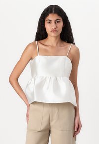 Hvit sateng peplum topp med tynne skulderstropper og innsamlet midje, flat front og jevn tekstur. Kombinert med beige bukser.