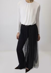 Witte lange mouwen top met een geplooide halslijn, gecombineerd met zwarte broek met gelaagde doorzichtige en kantaccenten. Puntige bruine hakken.