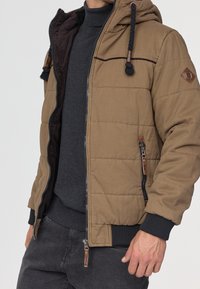 Tarnfarbene Pufferjacke mit Kapuze, dunkel gefüttert, mit Reißverschluss und Lederakzenten; gerippte Bündchen und Saum.