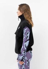 Schwarze Fleece-Weste mit hohem Kragen; getragen über einem langärmligen, lila und blauen gemusterten Oberteil. Das Design hat eine lockere Passform mit einem Reißverschlussdetail.