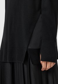 Pull en maille noire à manches longues avec fente latérale et manchette allongée sur une jupe plissée noire, main détendue sur le côté.
