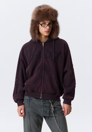 Ung person iført sorte briller med sorte kanter, brun pels hat, mørk lilla zip-up hættetrøje med stjerner på ærmerne og grå jeans med en sølvkæde hængende.