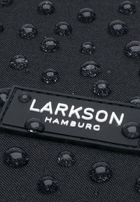 Svart strukturerat material med upphöjda vattendroppar, med en rektangulär svart etikett med "LARKSON HAMBURG" i vit text.