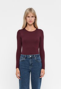 Donna con lunghi capelli biondi, che indossa un top aderente bordeaux a maniche lunghe e jeans blu a vita alta, in piedi davanti a uno sfondo bianco semplice.