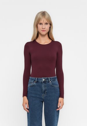 Kvinne med langt blondt hår iført en tettsittende burgunder langermet topp og høyt midje blå jeans, stående mot en ensfarget hvit bakgrunn.