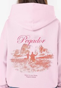 Rózsaszín kapucnis pulóver, amelyen egy nagy grafikás nyomat látható két figuráról egy táj előtt, rajta "Pegador" és "Térj vissza a gyökereidhez." felirat rózsaszínben.