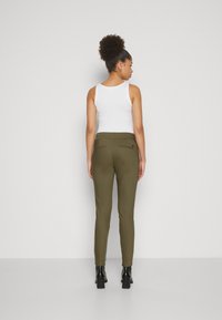 Olivově zelené kalhoty se slim fit, se dvěma zadními kapsami, v kombinaci s bílým tílkem a černými botami na vysokém podpatku.