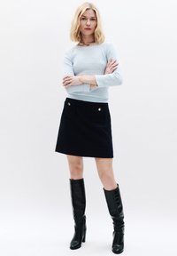 Helles blaues, geripptes Strickpullover mit langen Ärmeln und runden Halsausschnitt, kombiniert mit einem dunkelblauen A-Linien-Rock mit zwei vorderen Taschen und goldenen Akzenten. Schwarze kniehohe Stiefel runden das Outfit ab.