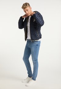 Giacca padded blu navy con cappuccio, chiusura a zip e tasche laterali; indossata sopra una t-shirt bianca e abbinata a jeans skinny azzurri e sneakers bianche.