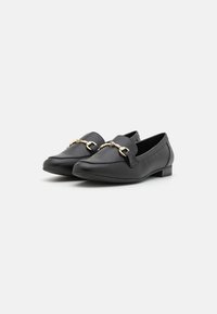 Mocassins en cuir noir avec un talon bas, présentant un accent de chaîne dorée à l'avant. Texture lisse, design à bout rond et coutures minimales.