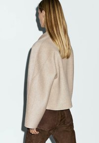 Beige wool jacket with a relaxed fit, dropped shoulders, and cropped hem, paired with brown suede pants. Textured fabric and minimal design.
