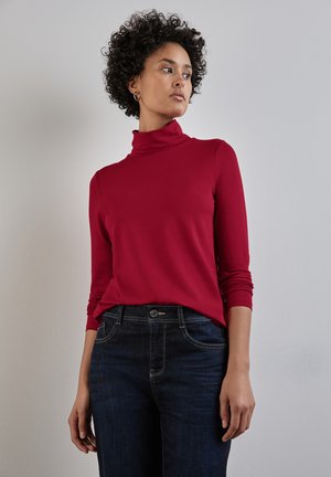 Rood coltrui met lange mouwen, gemaakt van een gladde stof, gecombineerd met donkerblauwe spijkerbroek. Kenmerkt zich door een strakke pasvorm en colneck.