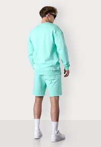 Sweatshirt vert menthe et shorts assortis en tissu doux, présentant une coupe décontractée et un petit logo dans le dos. Des baskets blanches complètent le look.