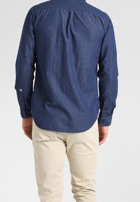 Mörkblå knäppskjorta med ett strukturerat mönster, långa ärmar och rundad nederkant. Matcher med ljusbeiga byxor.