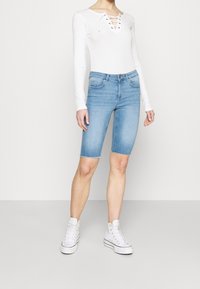 Bermudas de denim azul claro con dobladillo deshilachado, combinadas con una blusa de manga larga blanca con escote de cordones. Zapatillas blancas.