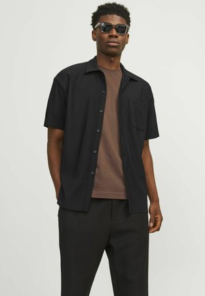 Short-sleeve black button-up shirt with a pocket, worn over a brown t-shirt, paired with black trousers and sunglasses, featuring a solid design.