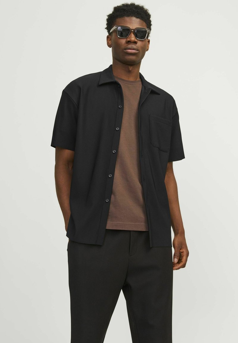 Short-sleeve black button-up shirt with a pocket, worn over a brown t-shirt, paired with black trousers and sunglasses, featuring a solid design.