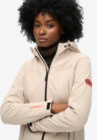 Beige softshell jack met een rits aan de voorkant, capuchon en geribbelde bruine kraag. Inclusief oranje Superdry-logo op de mouw en schouder.