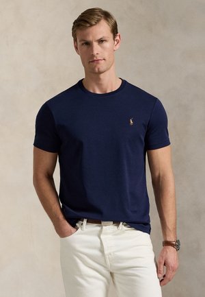 Férfi tengerészkék rövid ujjú pólóban, kis logóval, fehér nadrágban, barna övvel és barna karórával, egyszínű háttér előtt.