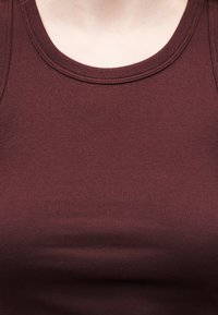 Gros plan sur une personne portant un débardeur côtelé bordeaux sans manches, montrant l'encolure et la partie supérieure de la poitrine.