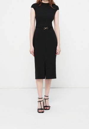 Femme portant une robe noire ajustée longueur genou avec des manches courtes, une petite ceinture et des sandales noires à talons ouverts, debout sur un sol blanc.