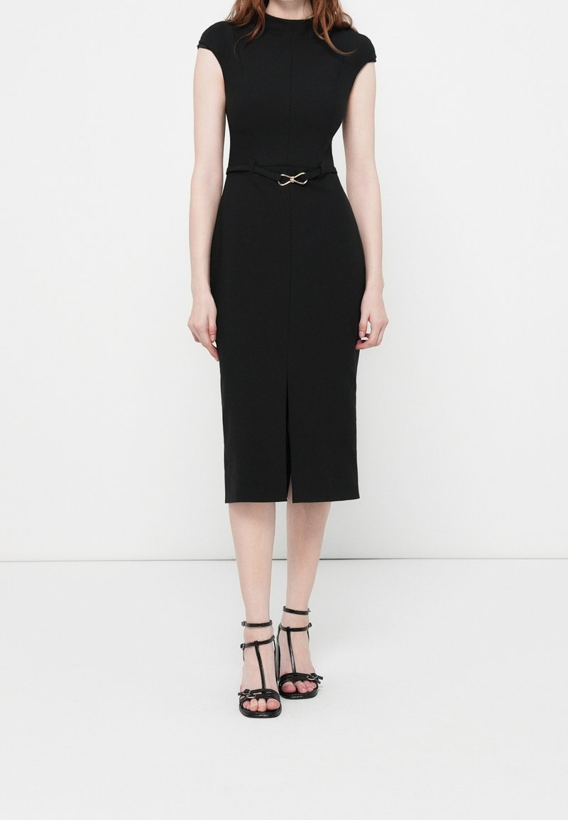 Femme portant une robe noire ajustée longueur genou avec des manches courtes, une petite ceinture et des sandales noires à talons ouverts, debout sur un sol blanc.