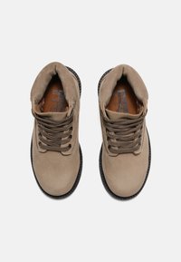 Stivaletti ankle in suede tan con lacci marroni, punta rotonda e suola in gomma nera texturizzata. L'interno presenta una fodera marrone e un dettaglio del logo.