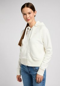 Weißer Reißverschluss-Hoodie mit Kapuze, großen Vordertaschen, gerippten Bündchen und glatter Textur. Kombiniert mit blauen Jeans.