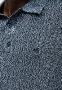 Gros plan d'un polo bleu chiné avec deux boutons et petit logo "MK" brodé sur la poitrine.