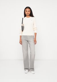 Kremowy sweter z splotem kablowym zestawiony z szarymi spodniami typu wide-leg i białymi trampkami. Modelka trzyma małą torebkę w wzory z paskiem na ramię.