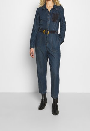 Femme portant une combinaison en denim foncé à manches longues, ceinture noire avec boucle ronde, et bottines noires, debout devant un fond uni.