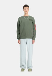 Felpa verde con collo a giro, tessuto testurizzato e una toppa sul petto. Abbinata a pantaloni in denim chiaro e scarpe da ginnastica con motivo verde.