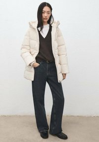 Jasnobeżowa kurtka puchowa z kapturem, noszona na ciemnobrązowym swetrze z dekoltem w serek, w połączeniu z luźnymi, ciemnymi dżinsami i czarnymi butami.