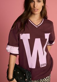 Mörk röd oversized sweatshirt med ett stort vitt "W" och texten "WICKED". Har kontrasterande ljusrosa ärmar och en v-ringning. Håller en svart nitarväska.