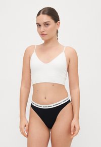 Bílý bralet s tenkými ramínky a designem ve tvaru V, doplněný černými tangy s bílým značkovým pasem.