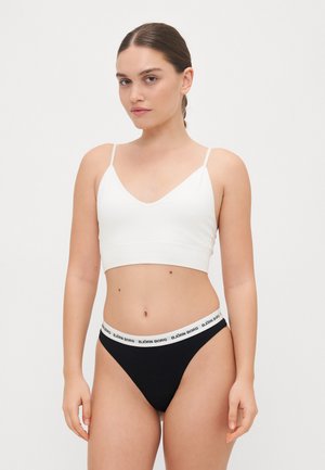 Hvit bralette med tynne stropper og V-halsdesign, paret med svart thong undertøy med en hvit merkebånd.