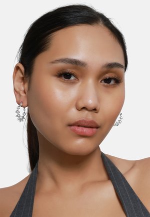 Silberne Creolenohrringe mit floralen Designdetails, die eine glänzende Oberfläche und eine strukturierte Textur aufweisen, getragen von einem Modell mit einem glatten Teint.