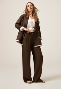 Bruin maatpak met brede pijpen en een blazer. Witte top eronder, met een gelaagde zoom. Lange ketting met ronde accenten.