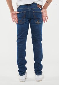 Jeans in denim blu con una vestibilità slim, dotati di due tasche posteriori e cuciture distintive. Indossati con sneakers bianche su uno sfondo semplice.