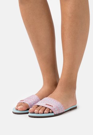 Pieds portant des sandales à enfiler avec des semelles bleues et des brides tressées rose pastel et blanches, se tenant sur une surface blanche.
