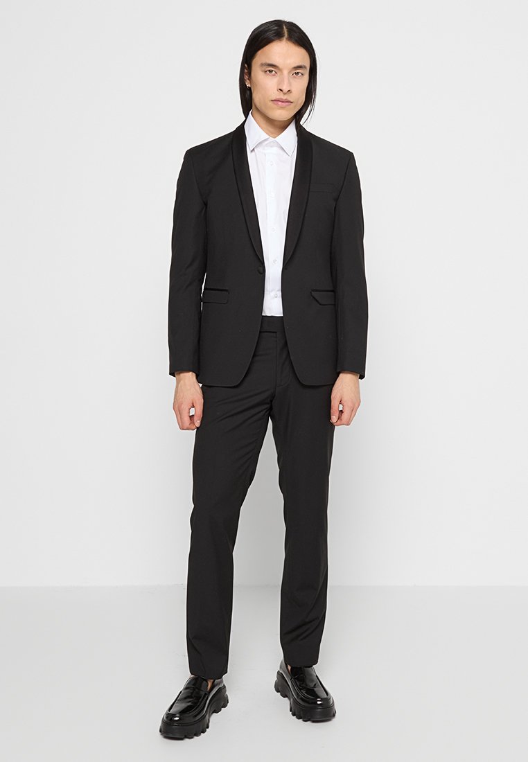 Costume noir avec un revers en satin, design ajusté et deux poches avant. Porté avec une chemise blanche à boutons et des chaussures noires brillantes.