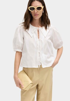 Kvinde iført hvid bluse med puffærmer, stor krave og bindebånd, beige bukser, lysegul clutch og cremefarvede katteøje-solbriller.