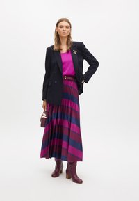 Blazer nero doppiopetto, top fucsia, gonna midi plissettata con righe verticali in bordeaux e blu navy, stivali con tacco marroni, borsa piccola.